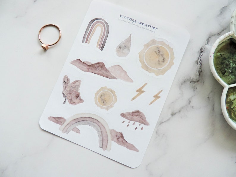 Puede incluir: Una hoja de pegatinas con tem&aacute;tica de clima vintage con ilustraciones de arco&iacute;ris, nubes, rayos, mariposas y soles en estilo acuarela. Las pegatinas est&aacute;n sobre un fondo blanco y tienen el texto "vintage weather" impreso en la parte superior.