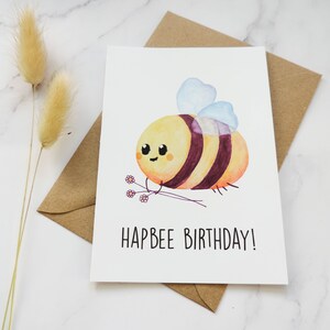 May include: A white card featuring a watercolour illustration of a cartoon bee holding a bunch of flowers. The text "Hapbee Birthday!" is printed below the bee.