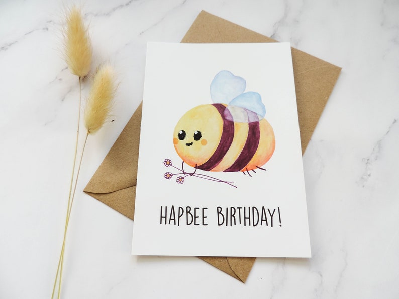 May include: A white card featuring a watercolour illustration of a cartoon bee holding a bunch of flowers. The text "Hapbee Birthday!" is printed below the bee.