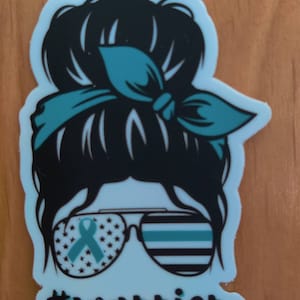 Puede incluir: Una pegatina azul turquesa y negra con la silueta de una mujer con un mo&ntilde;o desordenado, gafas de sol con una bandera estadounidense y una cinta azul turquesa en una lente y una bandera de l&iacute;nea azul fina en la otra lente. El texto #warrior est&aacute; debajo de las gafas de sol.