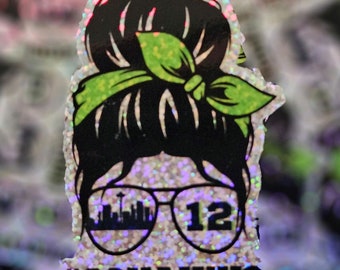 May include: A white glitter sticker featuring a black silhouette of a woman with a green bow in her hair. She is wearing sunglasses with a cityscape and the number 12. The text #GOHAWKS is below the sunglasses.