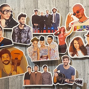 Op de afbeelding: Een verzameling uitgesneden stickers met illustraties van verschillende muzikanten en publieke figuren. De stickers tonen een verscheidenheid aan stijlen, met sommige die individuen in gedetailleerde portretten afbeelden en andere in meer gestileerde, grafische ontwerpen. De stickers zijn gerangschikt op een houten oppervlak.