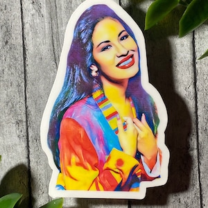 Op de afbeelding: Een kleurrijke sticker met een portret van Selena Quintanilla, een populaire zangeres, met lang donker haar en een brede glimlach. Ze draagt een veelkleurige jas met een gestreept patroon.