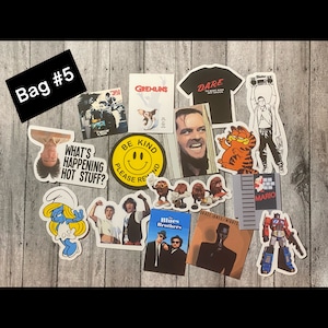 May include: A collection of stickers featuring various characters and designs, including Gremlins, the Blues Brothers, a smiling face with the words "Be Kind", a cartoon cat, a video game character, and a robot. The stickers are arranged on a wooden surface.