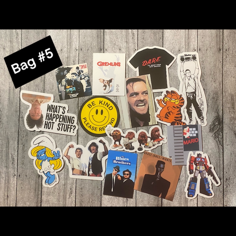 May include: A collection of stickers featuring various characters and designs, including Gremlins, the Blues Brothers, a smiling face with the words "Be Kind", a cartoon cat, a video game character, and a robot. The stickers are arranged on a wooden surface.