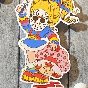 May include: A sticker featuring a cartoon character with a rainbow dress and a hockey mask holding a knife over another cartoon character with a strawberry dress and a red hat.