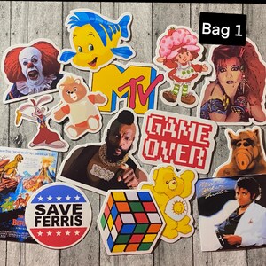 May include: A set of 11 colorful stickers featuring various characters and designs, including a cartoon clown, a mermaid, a Care Bear, a game over sign, a Rubik's Cube, and a Michael Jackson sticker. The stickers are arranged on a wooden surface with a white label that says "Bag 1".