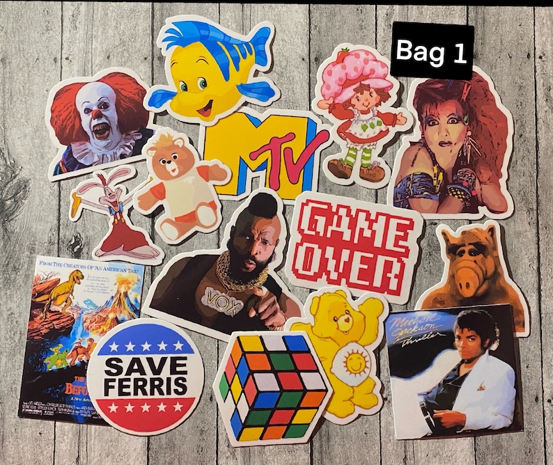 May include: A set of 11 colorful stickers featuring various characters and designs, including a cartoon clown, a mermaid, a Care Bear, a game over sign, a Rubik's Cube, and a Michael Jackson sticker. The stickers are arranged on a wooden surface with a white label that says "Bag 1".
