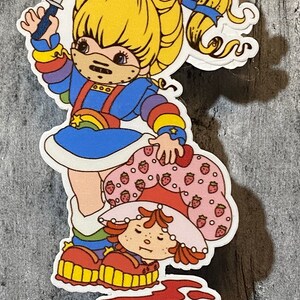 May include: A sticker featuring a cartoon character with rainbow-colored clothing holding a knife and standing over another cartoon character with a strawberry-patterned dress. The background is a wooden surface.
