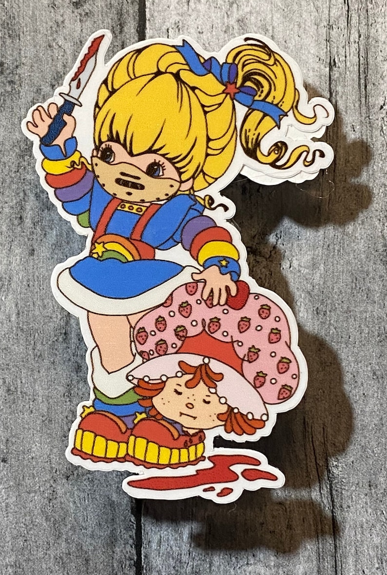May include: A sticker featuring a cartoon character with rainbow-colored clothing holding a knife and standing over another cartoon character with a strawberry-patterned dress. The background is a wooden surface.