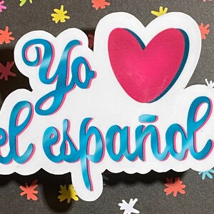 Op de afbeelding: Een witte sticker met de woorden "Yo el español" in turquoise script, een roze hart en een witte rand. De sticker ligt op een donkergrijze ondergrond met kleurrijke stervormige confetti.