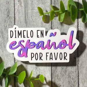 Op de afbeelding: Een witte sticker met de tekst "Dímelo en español por favor" in zwarte letters. Het woord "español" is in een paarse en roze gradiënt.