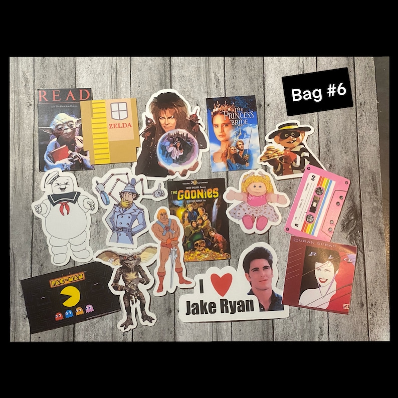 May include: A collection of stickers featuring various movie and pop culture characters, including the Ghostbusters, The Goonies, The Princess Bride, and Pac-Man. The stickers are arranged on a wooden surface and include text such as "READ", "ZELDA", "DURAN DURAN", and "Jake Ryan".