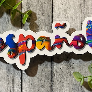 Può includere: Un adesivo bianco con la parola "Español" scritta in lettere colorate e stilizzate. Le lettere sono riempite con un motivo che ricorda un tessuto tradizionale tessuto.