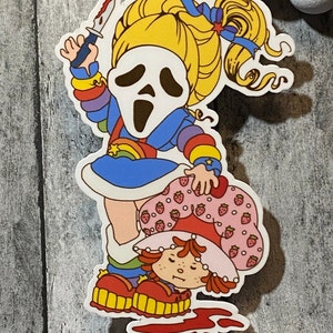 May include: A sticker featuring a cartoon character with a ghost mask holding a knife, standing on top of another cartoon character with a strawberry design. The background is a wooden surface.