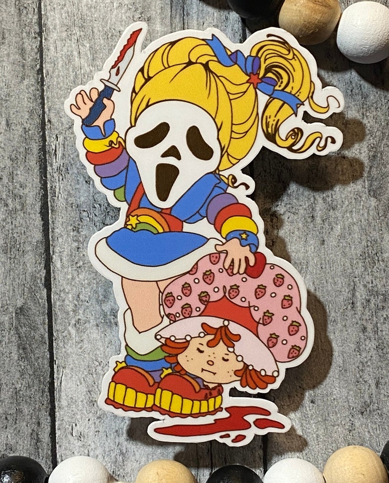 May include: A sticker featuring a cartoon character with a ghost mask holding a knife, standing on top of another cartoon character with a strawberry design. The background is a wooden surface.