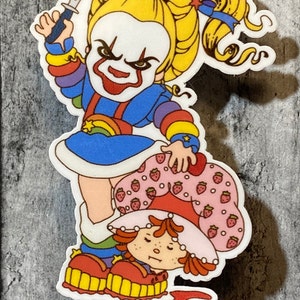 May include: A sticker featuring a rainbow-colored character holding a knife and standing over a strawberry-themed character. The rainbow character has a clown-like face with a red smile and a menacing expression. The strawberry character is lying on the ground with a pool of red liquid around them.