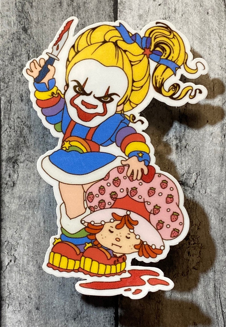May include: A sticker featuring a rainbow-colored character holding a knife and standing over a strawberry-themed character. The rainbow character has a clown-like face with a red smile and a menacing expression. The strawberry character is lying on the ground with a pool of red liquid around them.