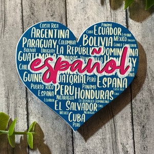 Op de afbeelding: Een blauw hartvormig bord met het woord "Español" in roze. Het bord is versierd met de namen van Spaanstalige landen, waaronder Argentinië, Bolivia, Colombia, Ecuador en Peru.