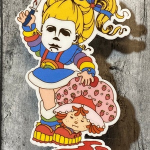 May include: A sticker featuring a cartoon character with a rainbow dress and a knife, standing over another cartoon character with a strawberry dress. The background is a wooden surface.