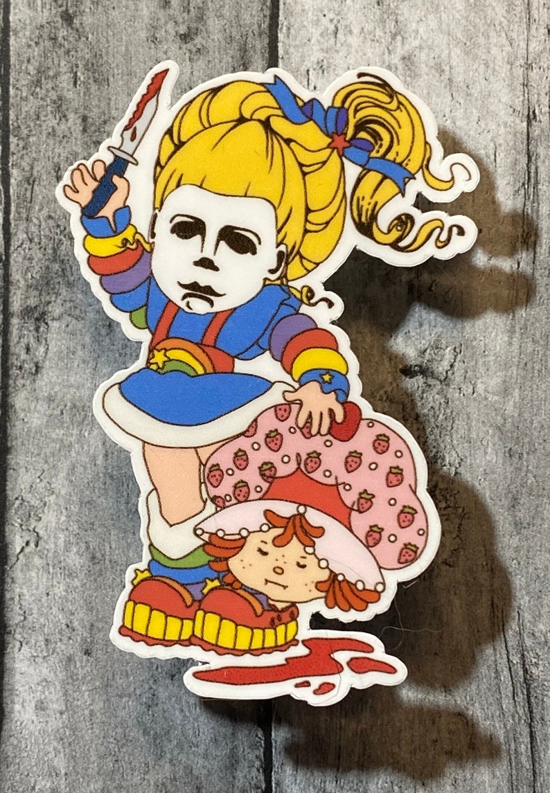 May include: A sticker featuring a cartoon character with a rainbow dress and a knife, standing over another cartoon character with a strawberry dress. The background is a wooden surface.