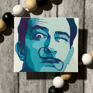 Op de afbeelding: Een canvas print met een gestileerd portret van een man met een opvallende snor. Het kunstwerk gebruikt tinten van teal, blauw en paars tegen een witte achtergrond. De uitdrukking van de man is intens, met één oog wijd open en het andere knipperend.