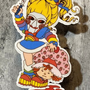 May include: A sticker featuring a cartoon character with a rainbow dress and a scary mask holding a knife over another cartoon character with a strawberry dress.