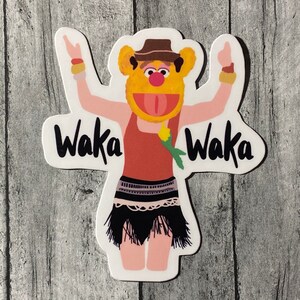 Op de afbeelding: Een sticker met een cartoonfiguur met oranje vacht, een rood-zwarte outfit en een bruine hoed. Het figuur houdt zijn armen in de lucht en de tekst "Waka Waka" staat aan beide kanten van het figuur.