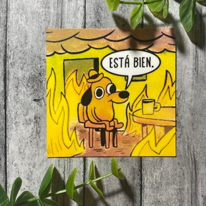 Op de afbeelding: Een gele hond met een hoed zit op een stoel in een kamer die in brand staat. De hond heeft een kalme uitdrukking en zegt "ESTÁ BIEN." in een spreekballon.