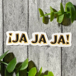 Op de afbeelding: Een witte sticker met zwarte tekst die "¡JA JA JA!" luidt. De tekst is geel omrand. De sticker bevindt zich op een grijze houten achtergrond.