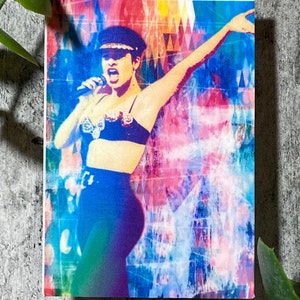 Op de afbeelding: Een levendige print met een zangeres in een zwarte outfit, inclusief een bh-top en een broek met hoge taille, met een bijpassende hoed. De achtergrond is een kleurrijk abstract ontwerp met tinten blauw, roze en geel. De zangeres houdt een microfoon vast en heeft een arm omhoog.