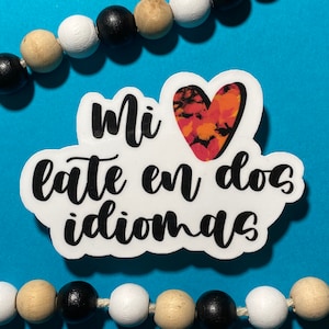 Op de afbeelding: Witte sticker met zwarte tekst die "Mi latte en dos idiomas" luidt, met een rood, oranje en zwart hart in de linkerbovenhoek.
