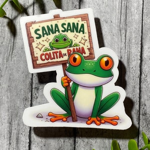 Op de afbeelding: Een groene kikker met oranje ogen houdt een houten bord vast met de tekst "Sana Sana Colita de Rana". De kikker zit op een witte achtergrond.