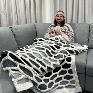 May include: A person wearing a grey and blue patterned sweater and a grey beanie sits on a grey couch with a large white and grey knitted blanket. The blanket has a geometric pattern.
