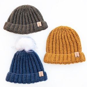 Puede incluir: Tres gorros de punto en diferentes colores: gris oscuro, amarillo mostaza y azul marino. Cada gorro tiene una pequeña etiqueta de cuero con el texto "Knits & Yarn" en ella. El gorro azul marino tiene un pompón blanco en la parte superior.