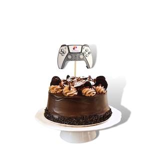 May include: A chocolate cake with chocolate frosting and sprinkles, topped with a white and gray video game controller cake topper.