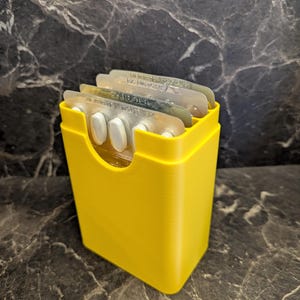 May include: A yellow pill organiser containing several blister packs of pills. The organiser is a small, rectangular container with a curved opening. The pills are white and visible.