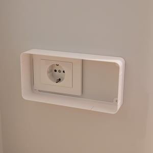 Könnte beinhalten: Eine weiße Steckdose mit zwei runden Löchern und einem Erdungsstift, montiert an einer hellgrauen Wand. Ein weißer rechteckiger Rahmen umgibt die Steckdose und sorgt für ein sauberes, modernes Aussehen. Die Steckdose ist für europäische Stecker ausgelegt.