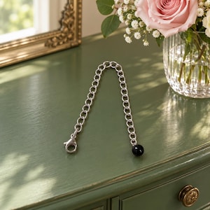 May include: A silver-toned metal chain bracelet featuring a series of oval-shaped links. One end of the chain is finished with a lobster claw clasp, while the other end is accented with a single, smooth, round, polished black bead.