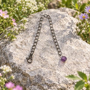 May include: A dark gunmetal chain extender featuring a series of interlocking oval links, a lobster claw clasp at one end, and a single round, polished purple amethyst bead dangling from the opposite end of the metal chain.