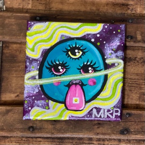 May include: A hand-painted canvas featuring a blue, cartoon-like planet with three eyes, eyelashes, and a pink tongue sticking out. The planet is surrounded by a yellow ring and a purple and white starry background. The artist's signature "MRP" is in the bottom right corner.