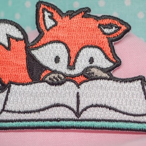 Puede incluir: Parche bordado con un zorro de dibujos animados con pelaje naranja, cara blanca y detalles negros, leyendo un libro. El libro tiene páginas blancas y un borde turquesa. El parche está sobre un fondo de tela rosa.