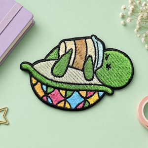Puede incluir: Un parche bordado que muestra una tortuga verde acostada boca arriba mientras sostiene una taza de café de color canela y blanco. La tortuga tiene un caparazón colorido con un patrón geométrico de diamantes rosas, azules y amarillos.
