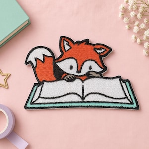 May include: An embroidered patch featuring a stylised orange and white fox with black outlines, depicted reading an open white book with a teal border. The fox has a bushy tail, pointed ears, and small paws resting on the pages of the open book.