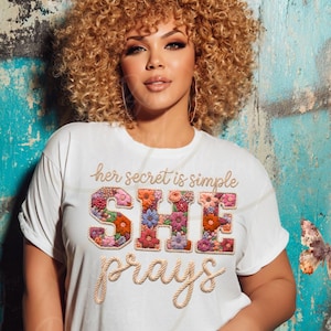 Könnte beinhalten: Weißes T-Shirt mit dem Text "her secret is simple SHE prays". Das Wort "SHE" ist mit floralen Mustern gestaltet. Das T-Shirt wird von einer Person mit lockigem Haar getragen.