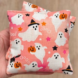 May include: A square fabric bean bag featuring a repeating pattern of white ghosts, orange pumpkins, and black stars on a light pink background. Some ghosts wear black witch hats, and the word Boo is printed in white among the festive icons.