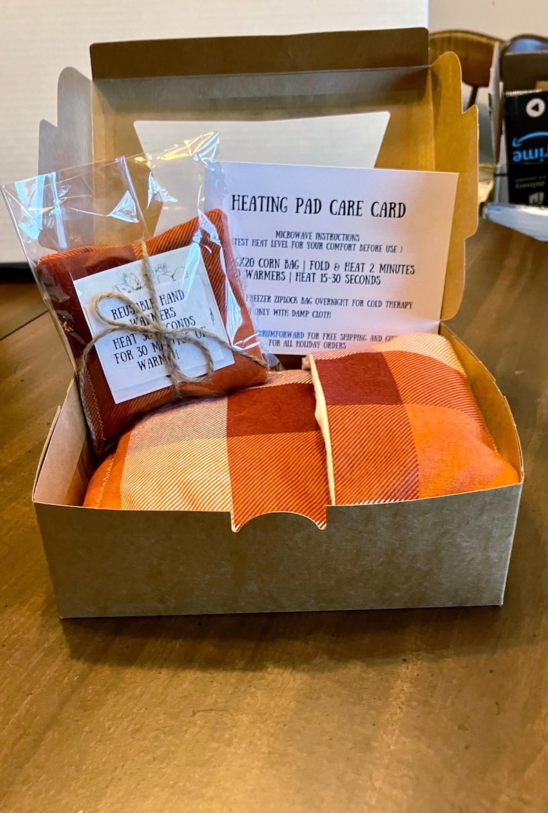 May include: A brown cardboard box with a plaid fabric and two red fabric pouches inside. The box has a label that reads "Heating Pad Care Card" with instructions for using the heating pad. The label also says "Reusable Hand Warmers" and "Heat 30 Seconds for 30 Minutes Warm".