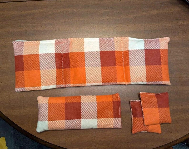 May include: Four orange and white plaid fabric heat packs. The largest pack is rectangular and divided into three sections. The other three packs are smaller and square.
