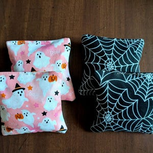 May include: Four small, square fabric pouches. Two pouches have a pink background with white ghosts, orange pumpkins, and black stars. The other two pouches have a black background with a white spiderweb design.