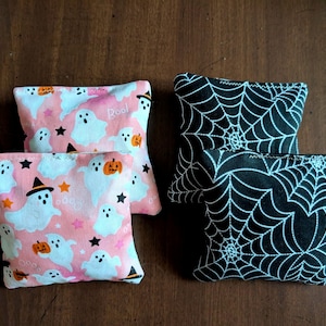 May include: Four small, square fabric pouches. Two pouches feature a pink background with white ghosts, orange pumpkins, and black stars. The other two have a black background with a white spiderweb design.